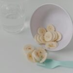 Sliced banana pieces in a white bowl with a mint fork and glass of water on a clean table.