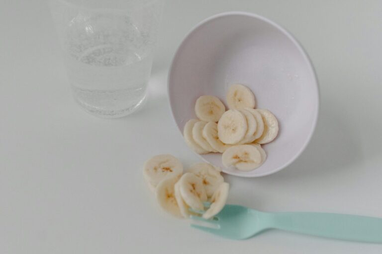 Sliced banana pieces in a white bowl with a mint fork and glass of water on a clean table.